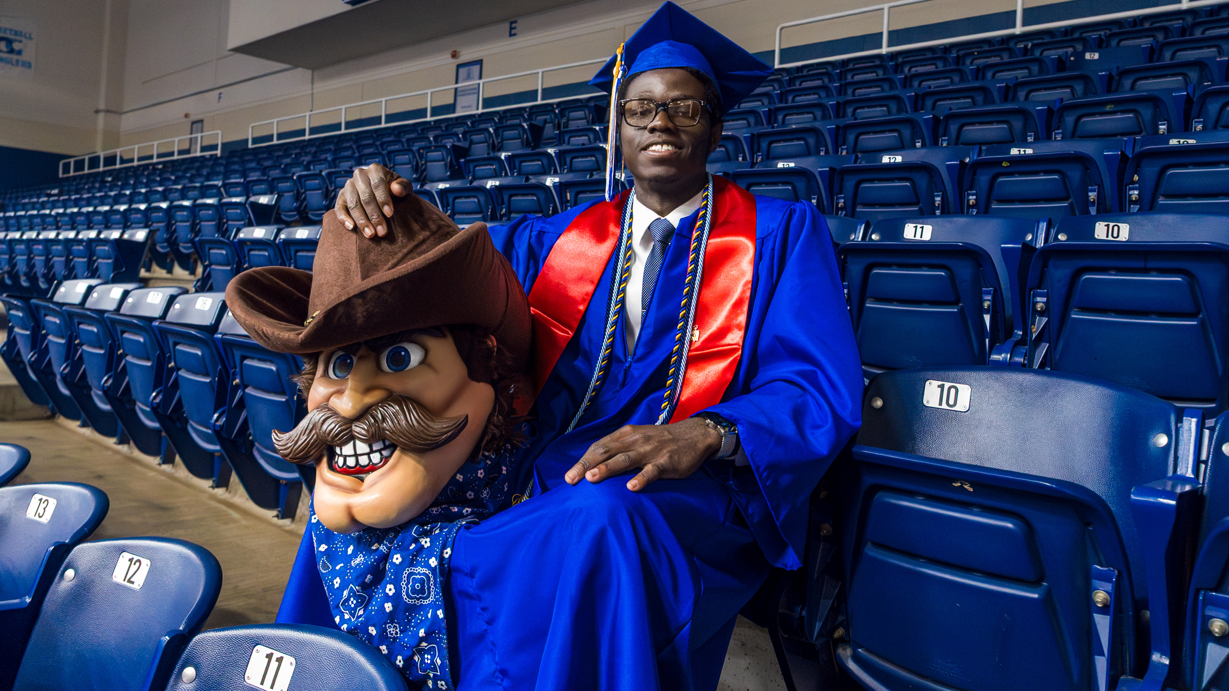 A smiling graduate in a blue cap and gown sits in stadium seating, resting his hand on a large mascot head with a cowboy hat and mustache.