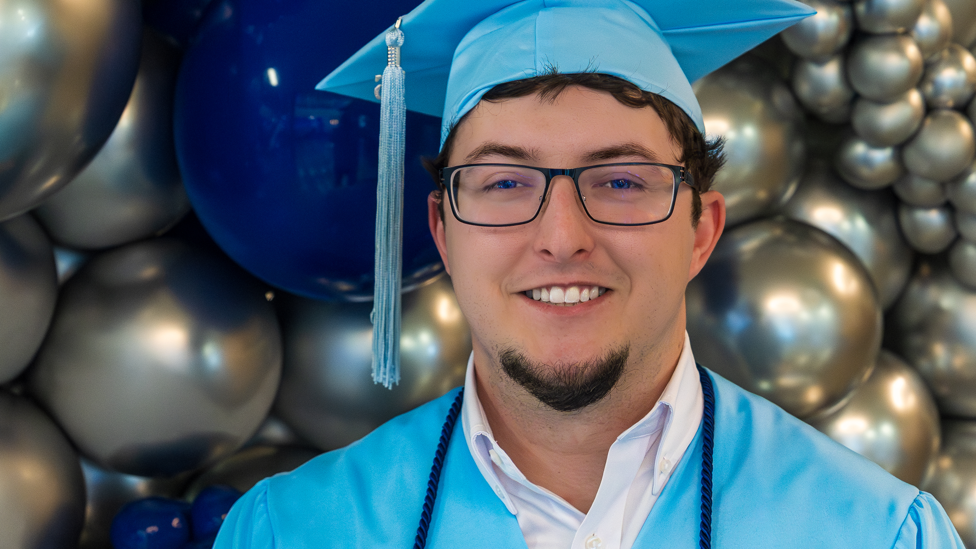 A smiling young man with glasses wearing a light blue graduation cap and gown in front of a blue and silver balloon backdrop.