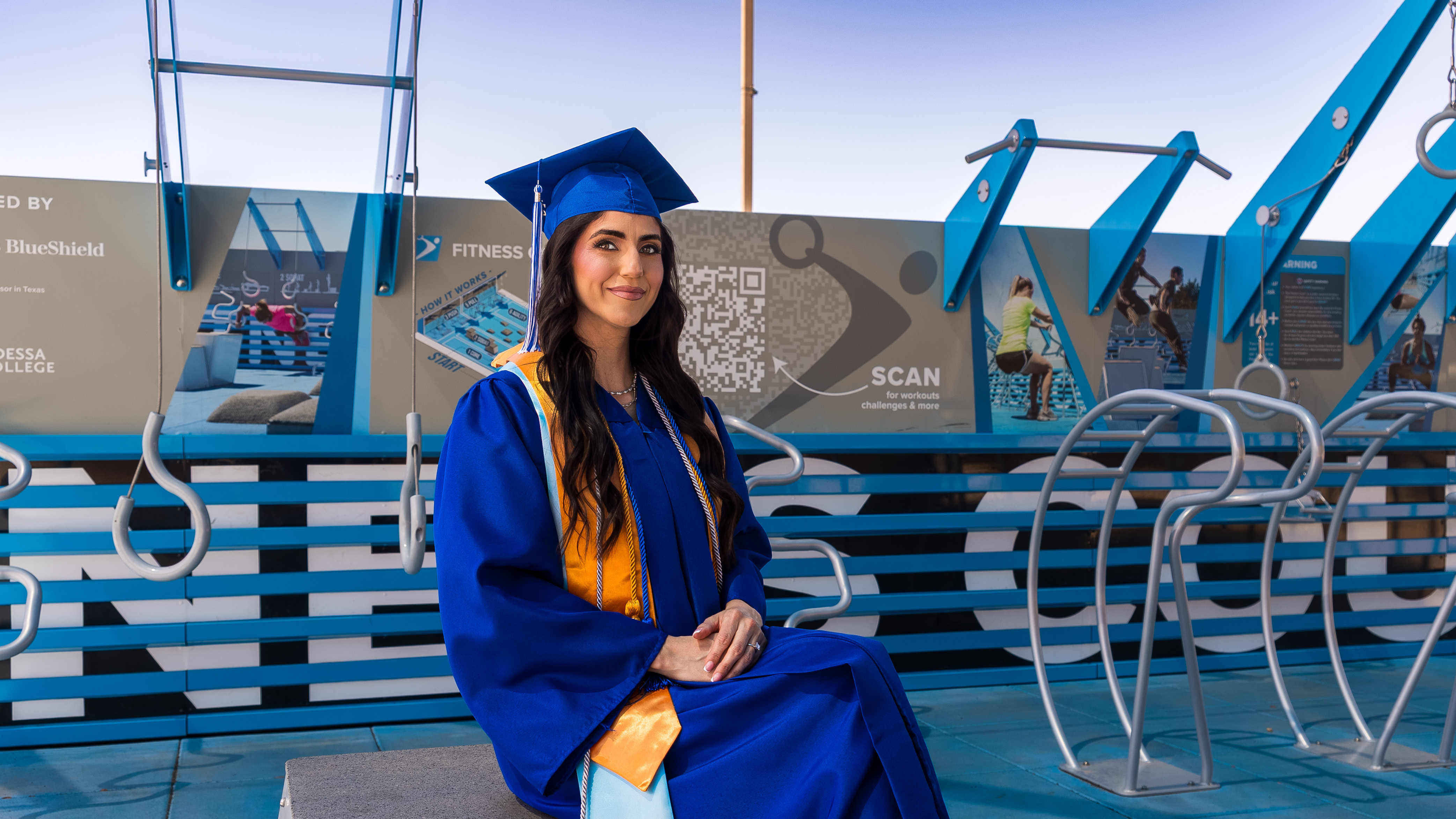 A young woman in a blue graduation cap and gown sits at an outdoor fitness park, wearing a gold and light blue stole with academic cords.