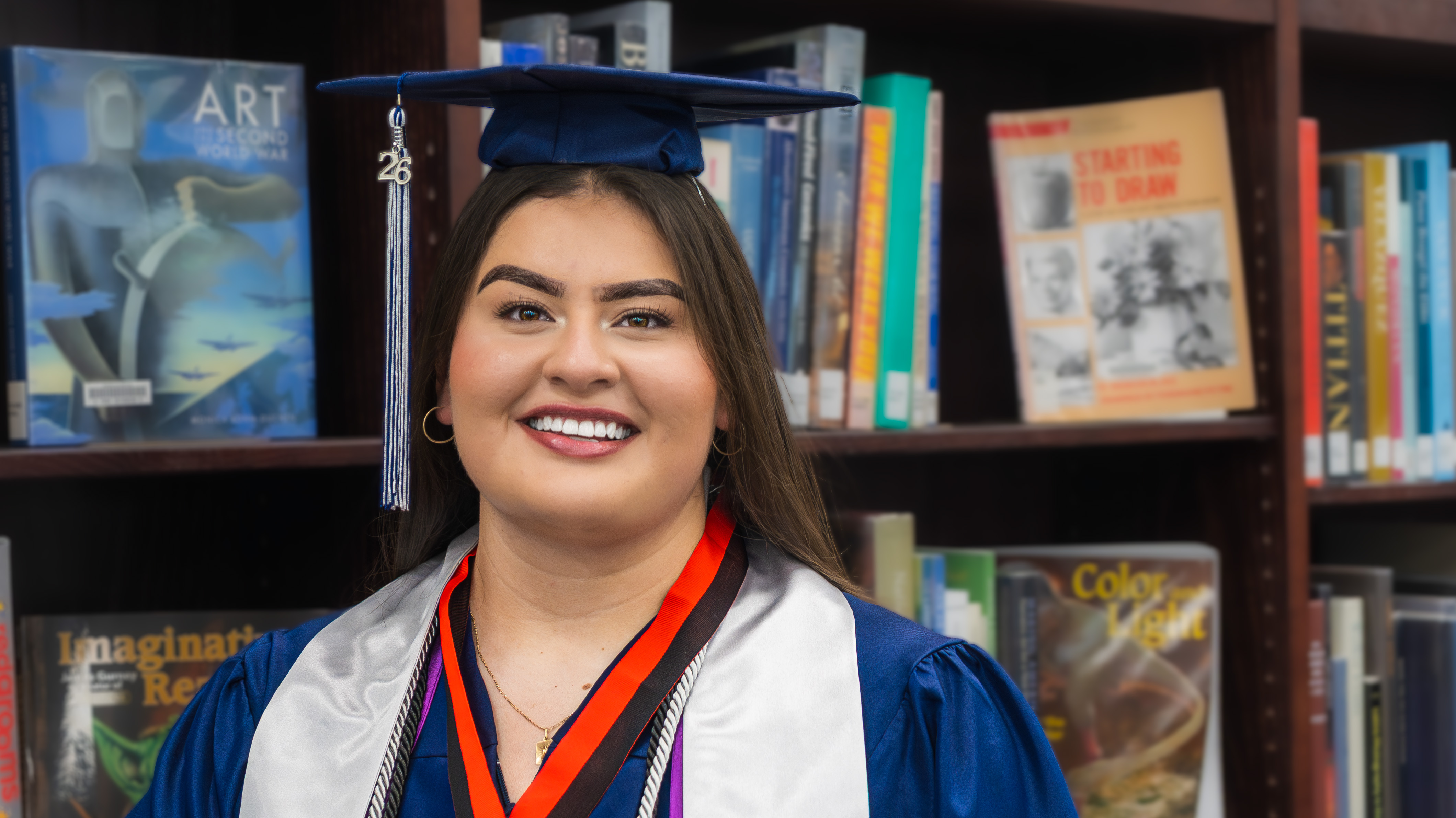 A smiling young woman in a navy graduation cap and gown with a silver stole, posing in front of a bookshelf filled with books.