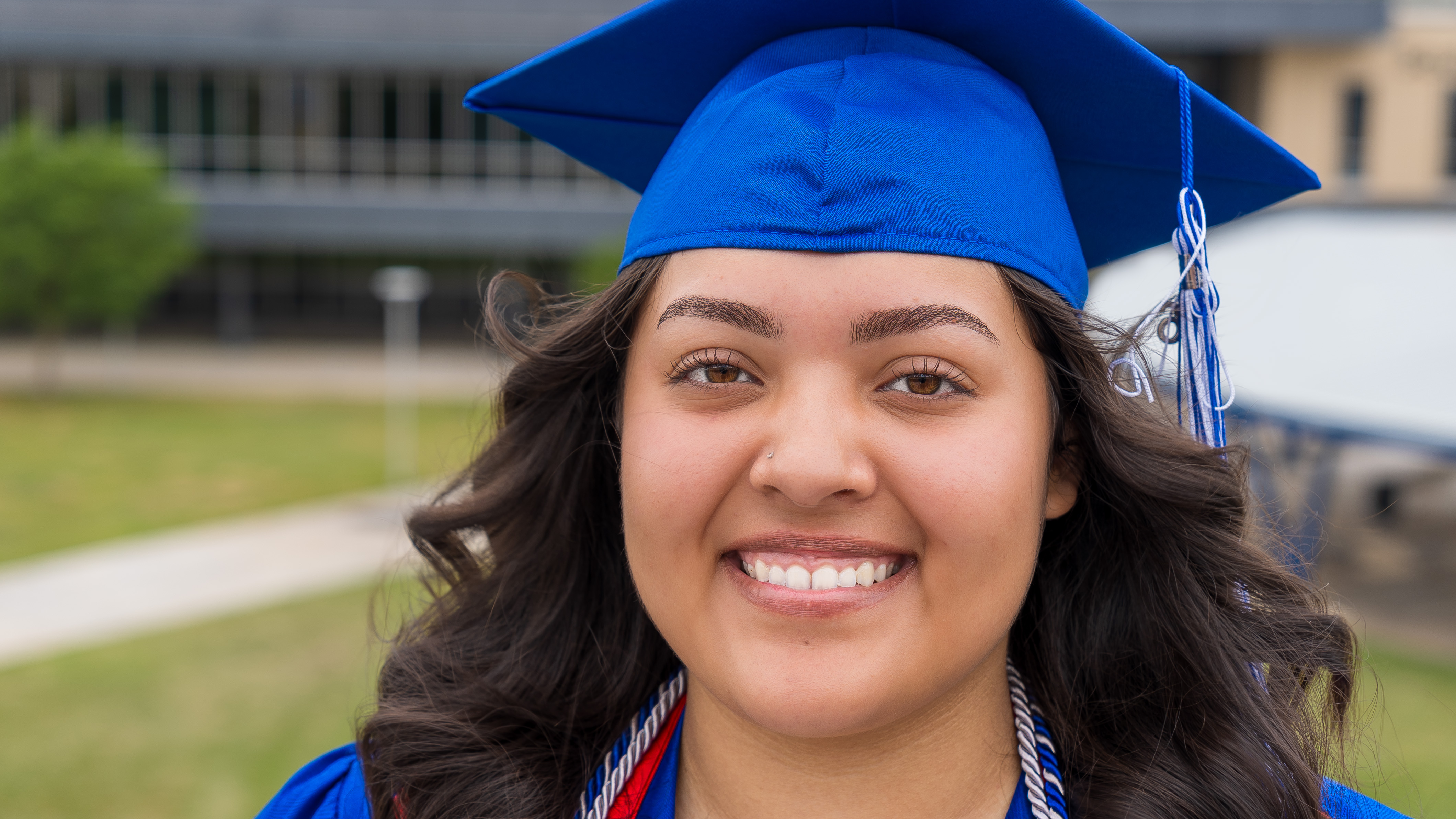 A close-up, smiling portrait of a young woman with long wavy hair wearing a bright blue graduation cap and gown with academic cords.