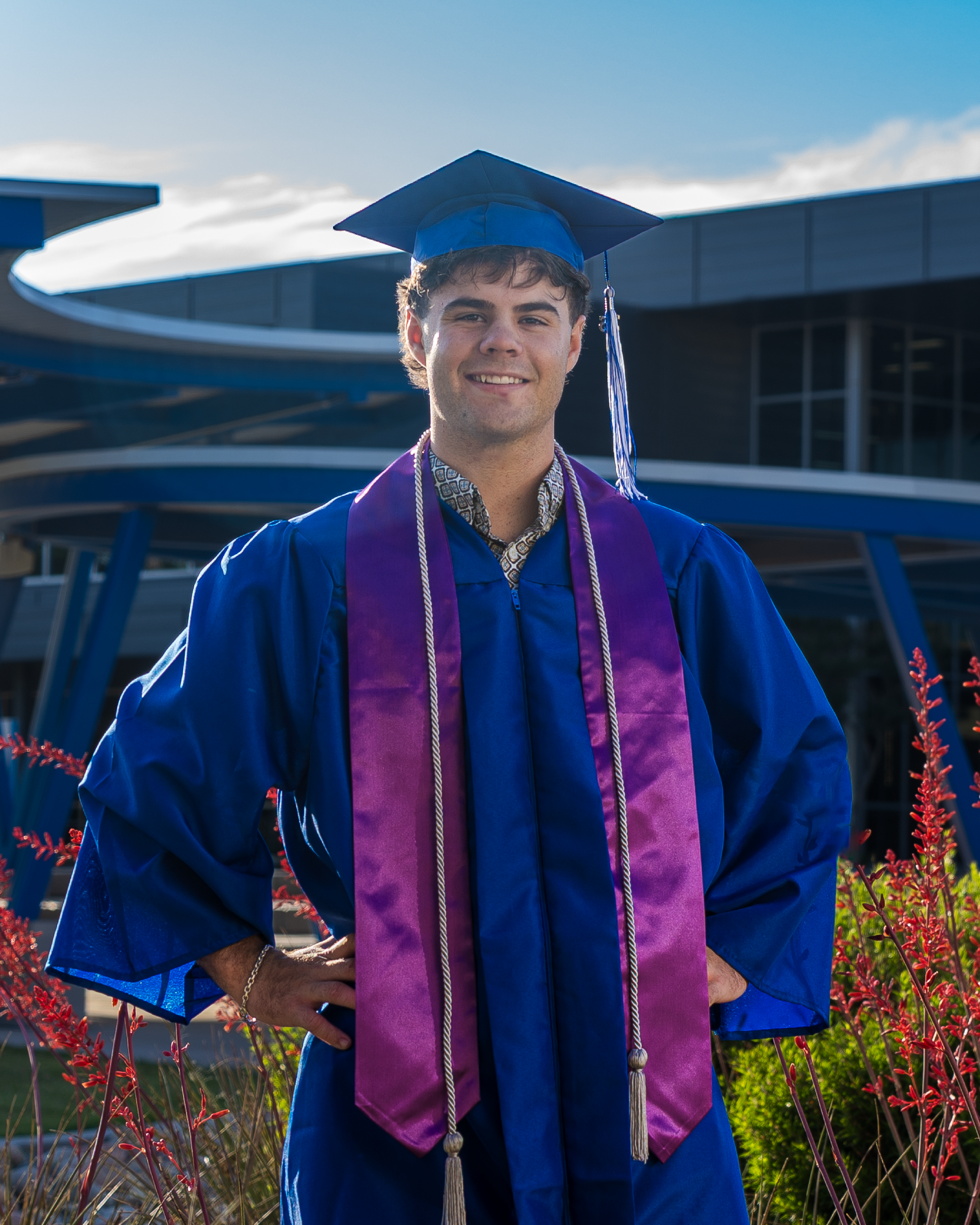 A smiling graduate stands outdoors wearing a blue cap and gown with a purple stole and honor cords, posing with hands on hips. He is in front of a modern campus building with curved architecture, with red flowering plants and greenery in the foreground under a bright sky.