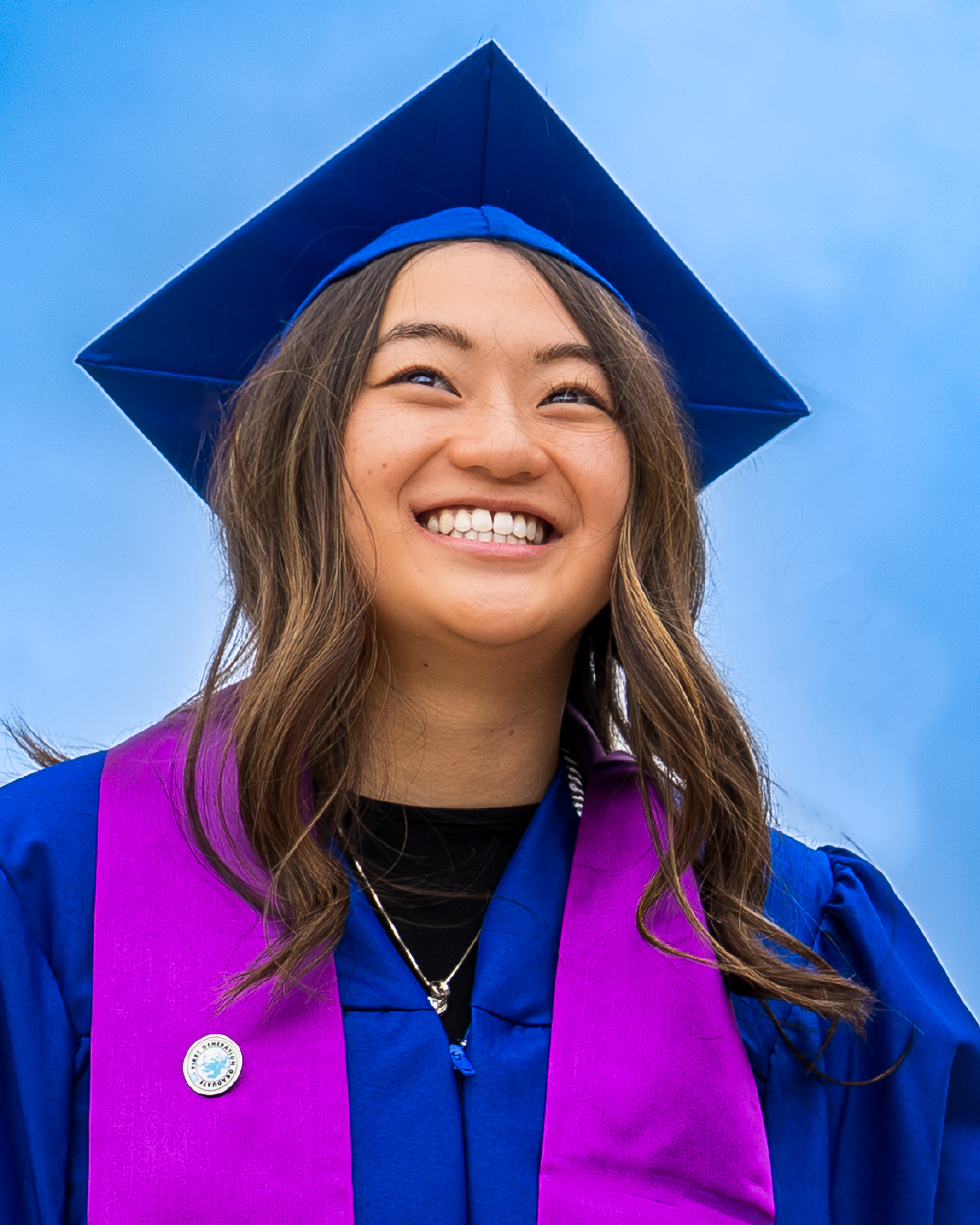 A smiling young woman wearing a blue graduation cap and gown with a purple stole against a bright blue sky.
