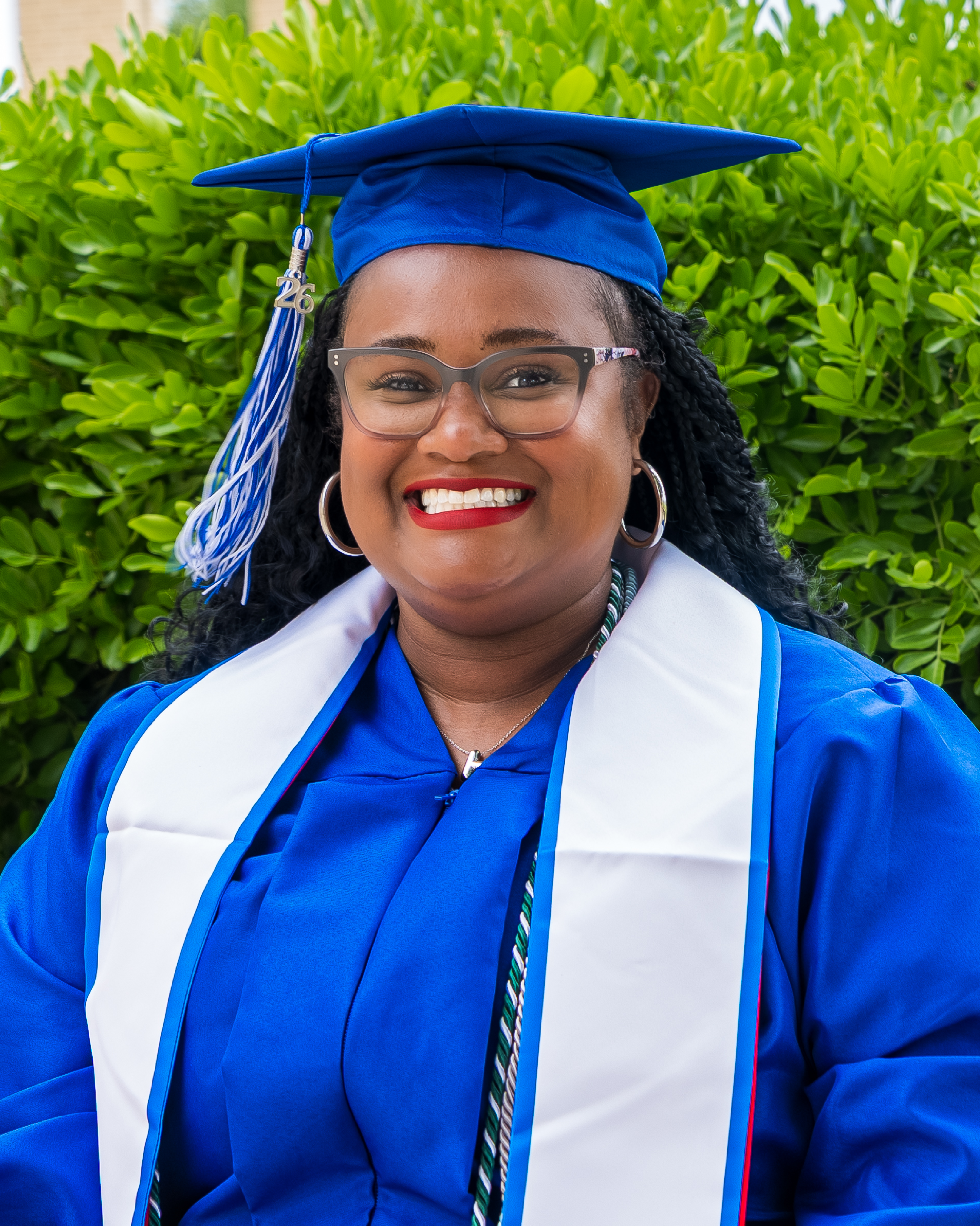 A smiling graduate wearing a bright blue cap and gown with a white stole poses outdoors in front of green bushes. She has long dark braided hair, wears glasses and hoop earrings, and her graduation cap features a blue and white tassel with a “26” charm.