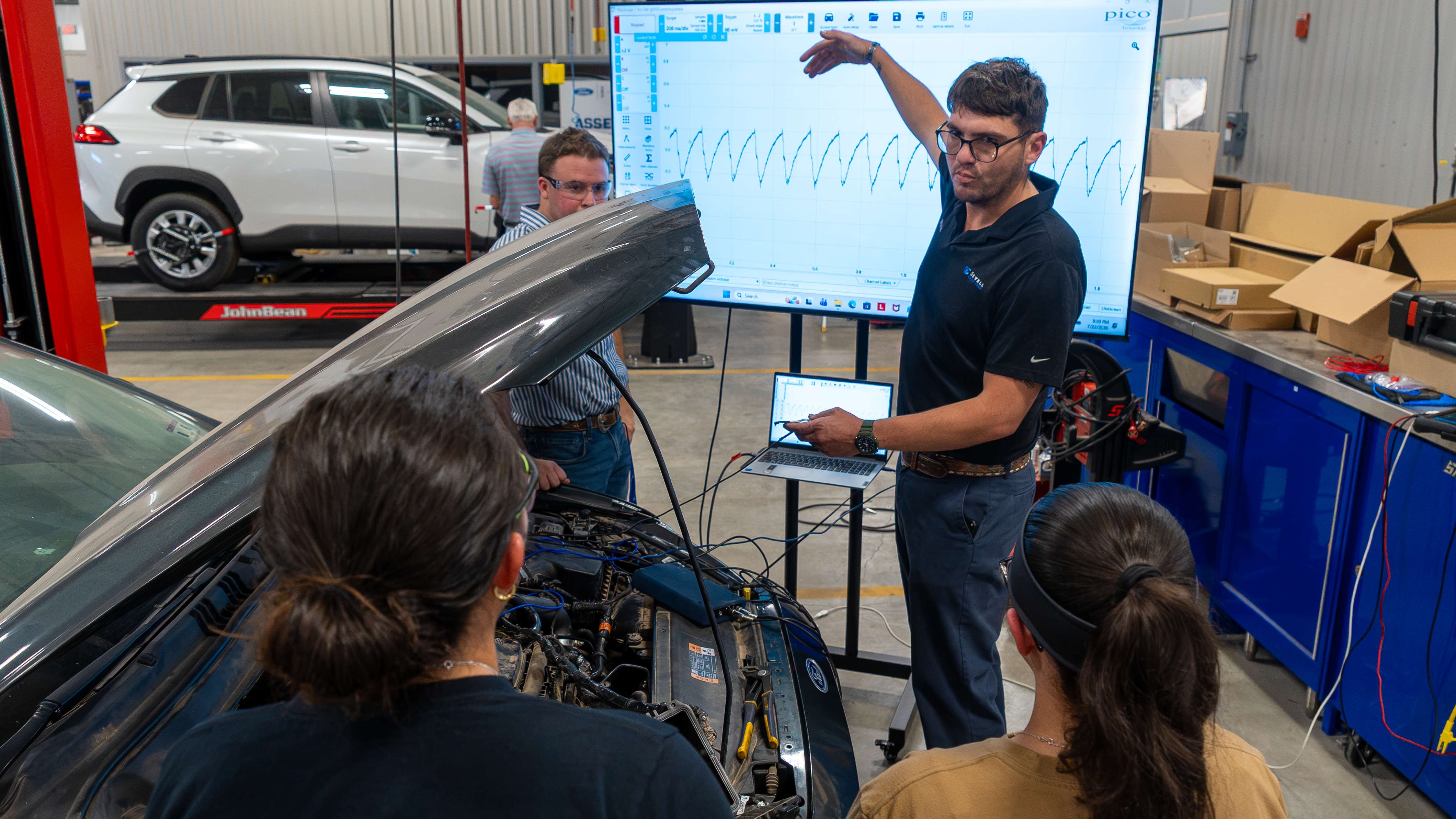 A professor teaching in front of a car with his students around
